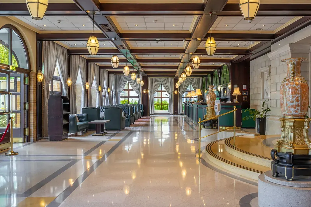 Az Ensana Grand Margitsziget patinás lobby-ja, ahol márvány padló, elegáns csillár, zöld bársony fotel és arany váza látható panorámaablakokkal