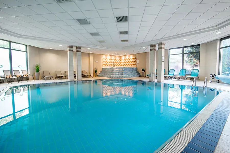A Rubin Wellness & Conference Hotel Budapest fedett élménymedencéje, ahol türkiz víz, fehér oszlopok és mozaikdíszes lépcső látható nyugágyakkal